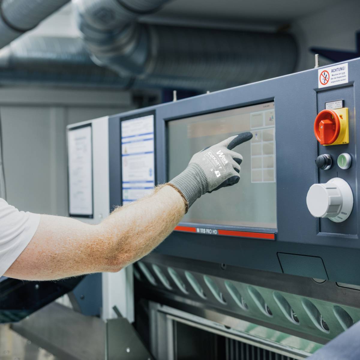 Eine Person trägt Handschuhe und bedient ein Touchscreen-Panel an Industriemaschinen in einer Anlage und passt die Einstellungen während eines Arbeitsprozesses an.