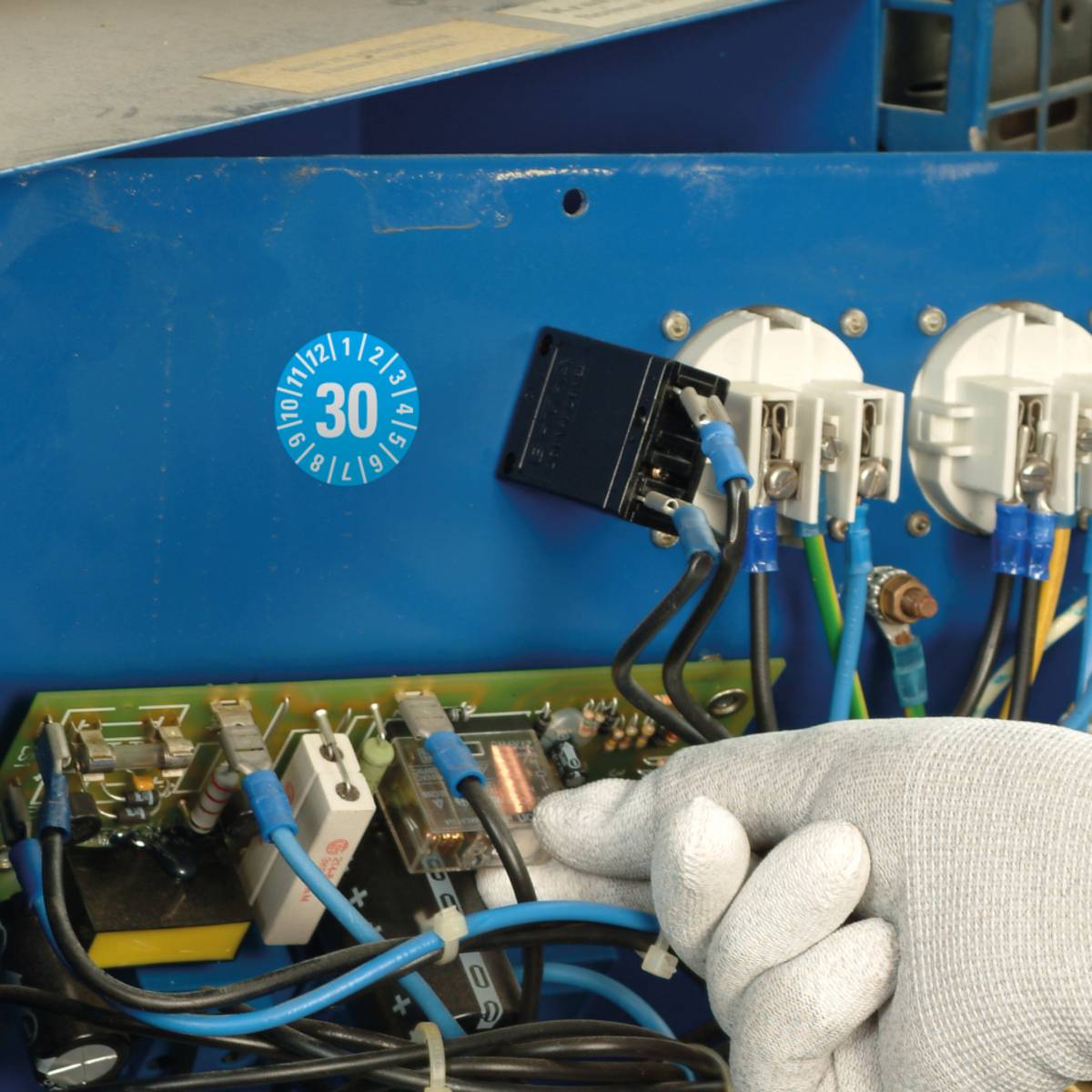 Eine behandschuhte Hand arbeitet an elektrischen Leitungen, die mit einer Leiterplatte und Stromsteckdosen verbunden sind. Ein blauer Inspektionsaufkleber mit der Markierung „30