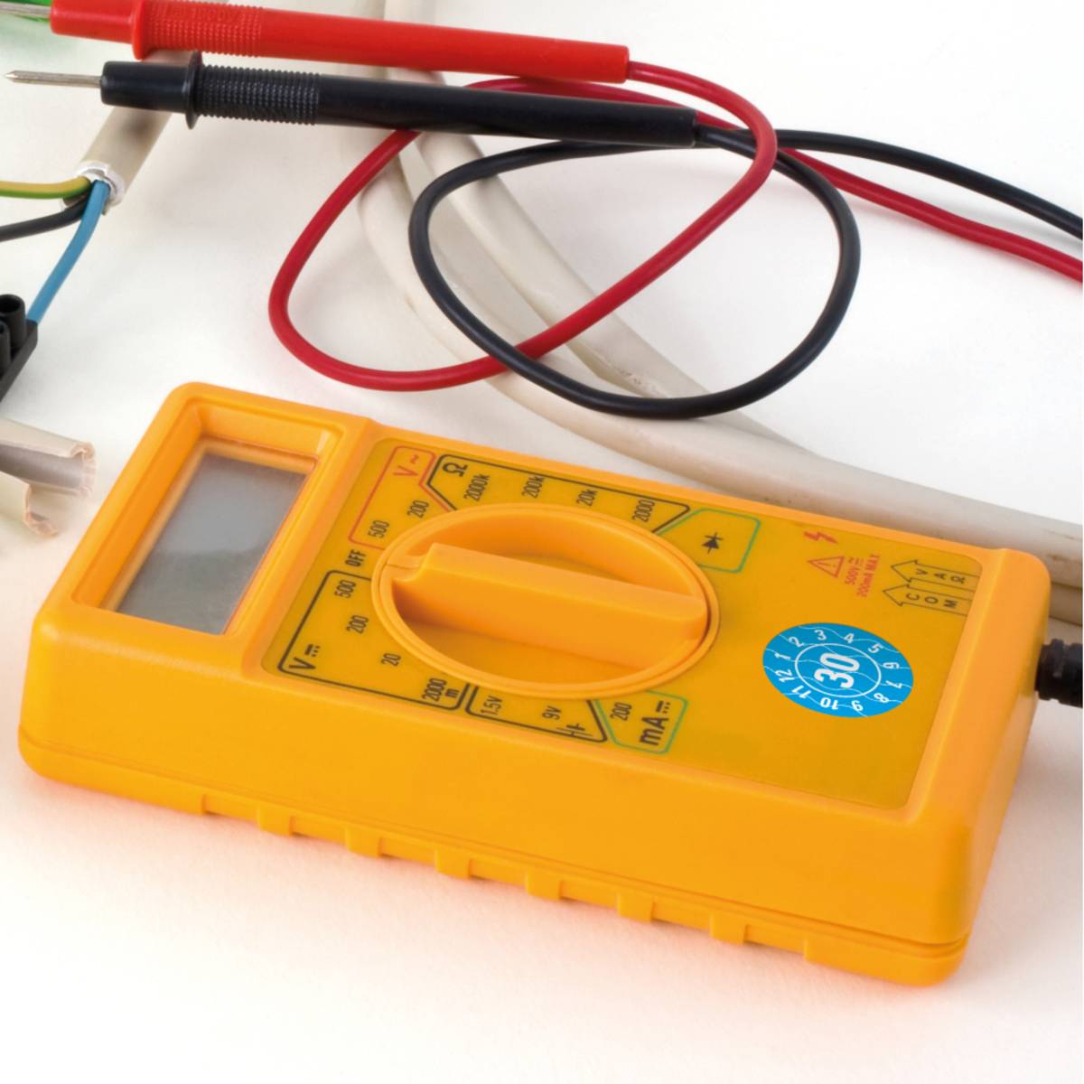 Ein gelbes Multimeter mit angebrachten roten und schwarzen Messleitungen ist auf Spannungsmessung eingestellt und auf einer weißen Oberfläche platziert, mit Drähten in der Nähe.