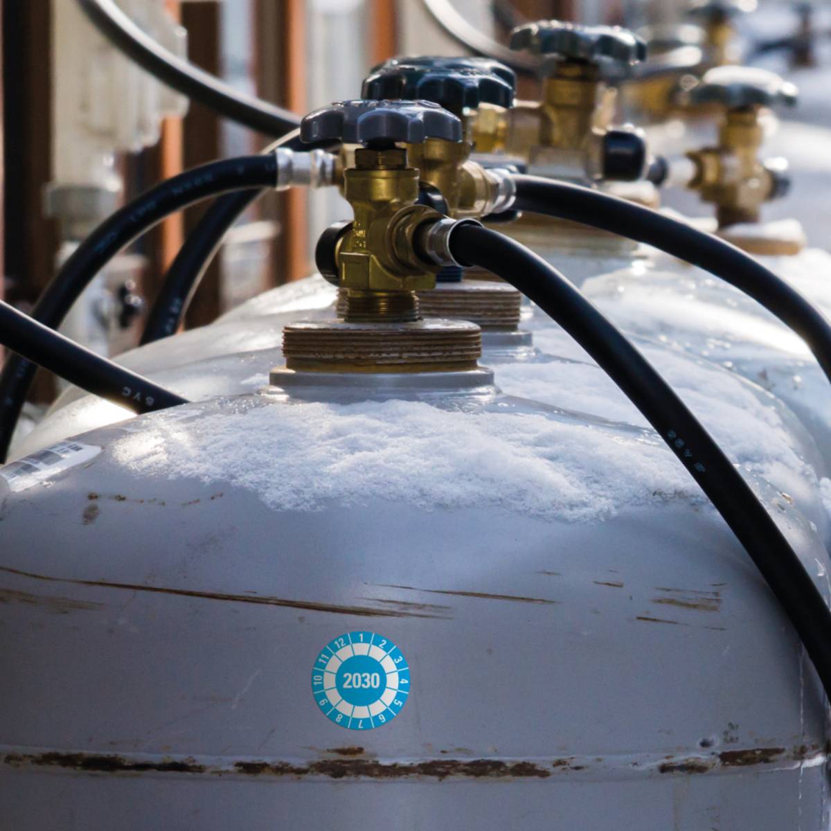 Eine Reihe großer Metalltanks, die durch schwarze Schläuche verbunden sind, mit Reifbildung auf der Oberfläche. Ein kleiner blauer Aufkleber mit der Aufschrift „2030