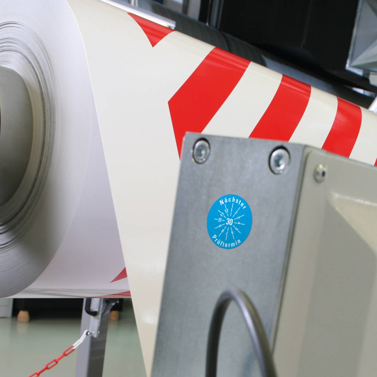 Eine große industrielle Rolle weißen Papiers mit roten Streifen ist auf einer Maschine montiert. Ein kreisförmiges blaues Etikett auf der Maschine liest „Machine Performance