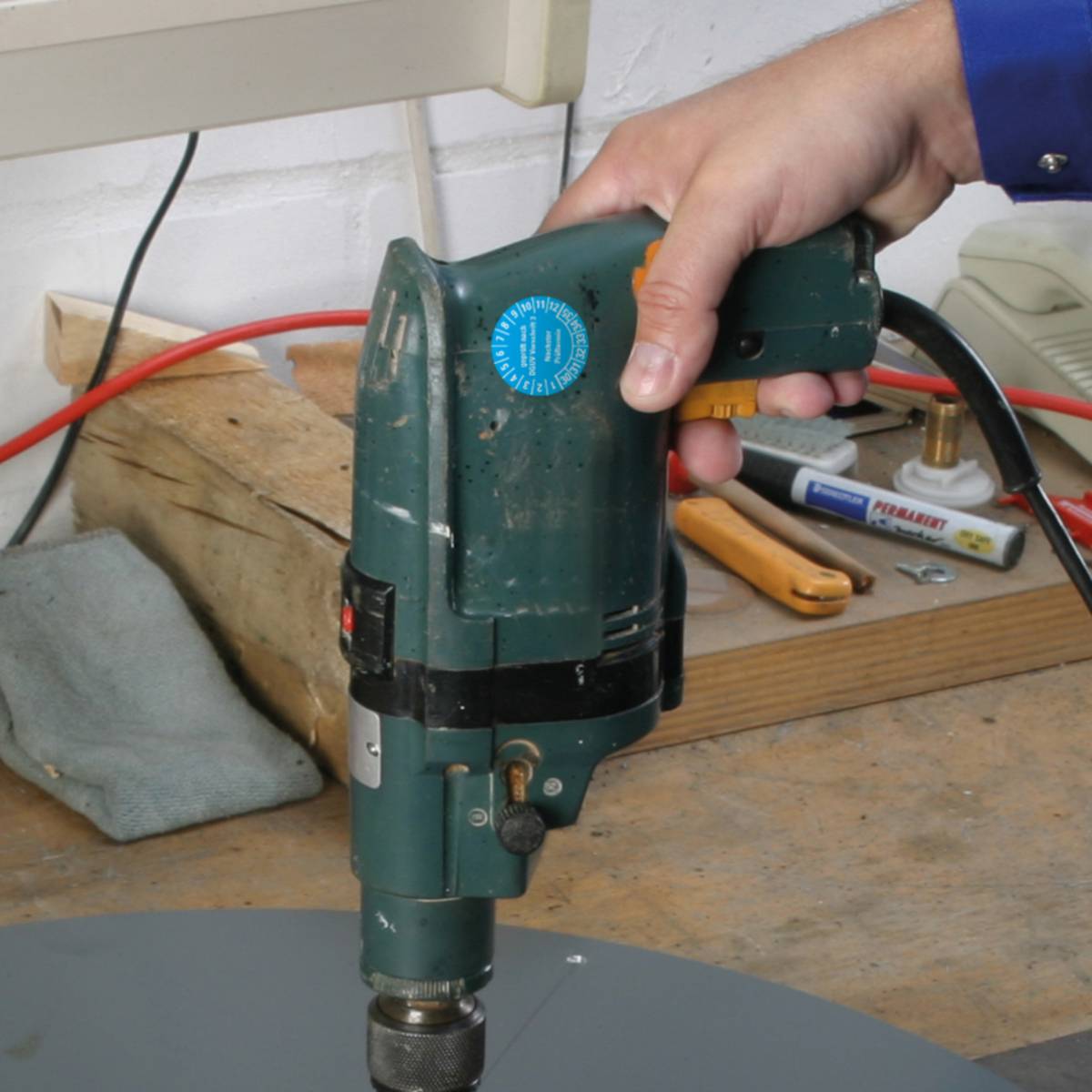 Eine Person, die einen grünen Elektroschrauber auf einer Holzwerkbank benutzt, umgeben von verschiedenen Werkzeugen und einer grauen Oberfläche.