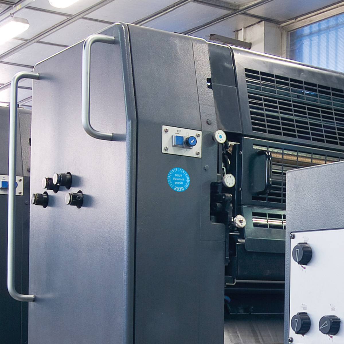 Eine große Industrialdruckmaschine in einer Fabrikumgebung mit Metallpaneelen, Steuertasten und einem teilweise sichtbaren Papierzuführungssystem.