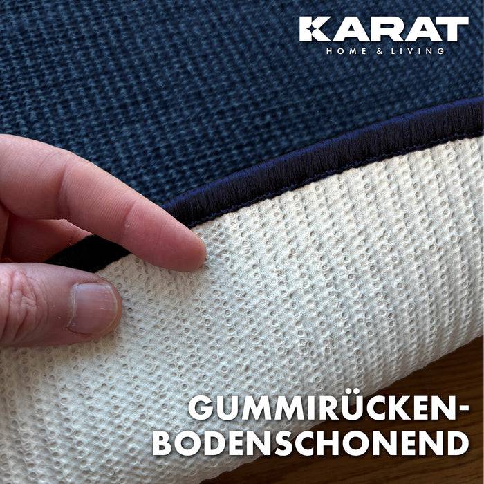 Eine Hand hebt einen dunkelblauem Teppich an und enthüllt dessen strukturierte, weiße Gummirückseite mit der Aufschrift 'Gummirücken-Bodenschonend' und 'KARAT Home & Living'.