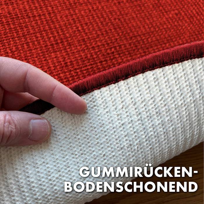 Eine Hand hebt eine Teppichkante an und zeigt die rutschfeste Unterseite sowie die Wörter 'Gummirücken-Bodenschonend'. Der Teppich ist rot.