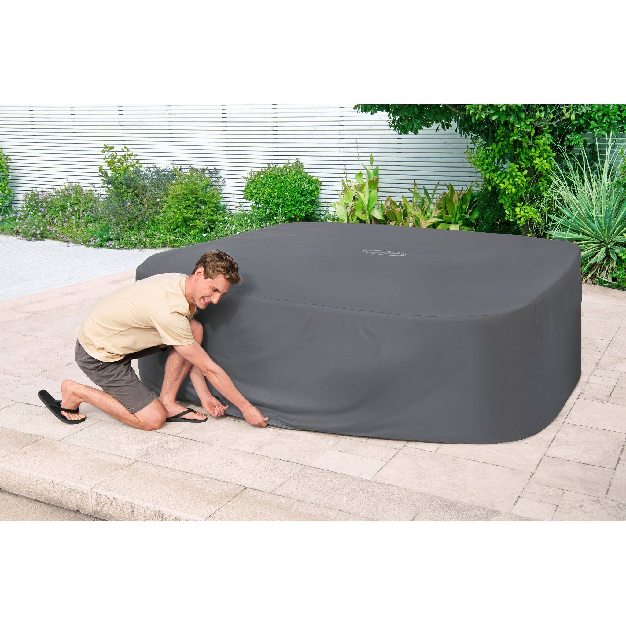 Eine Person hockt sich hin, um eine große graue Abdeckung auf einem Außenwhirlpool anzupassen, der auf einer Steinterrasse steht und von grünen Pflanzen umgeben ist.