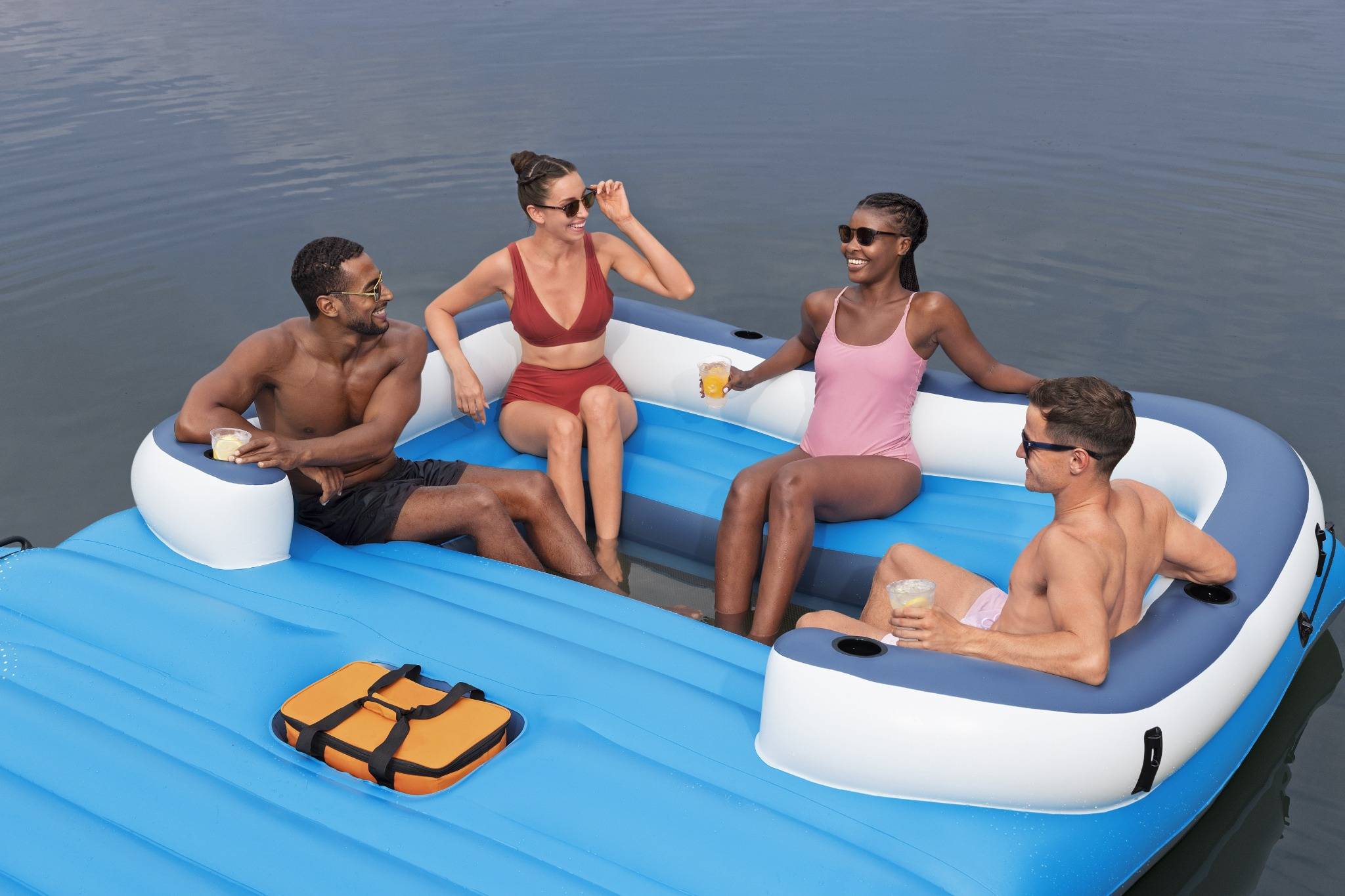 Vier Menschen in Badekleidung entspannen sich auf einem aufblasbaren Floß in einem See, lächeln und genießen Getränke, mit einer Kühlbox in der Nähe.