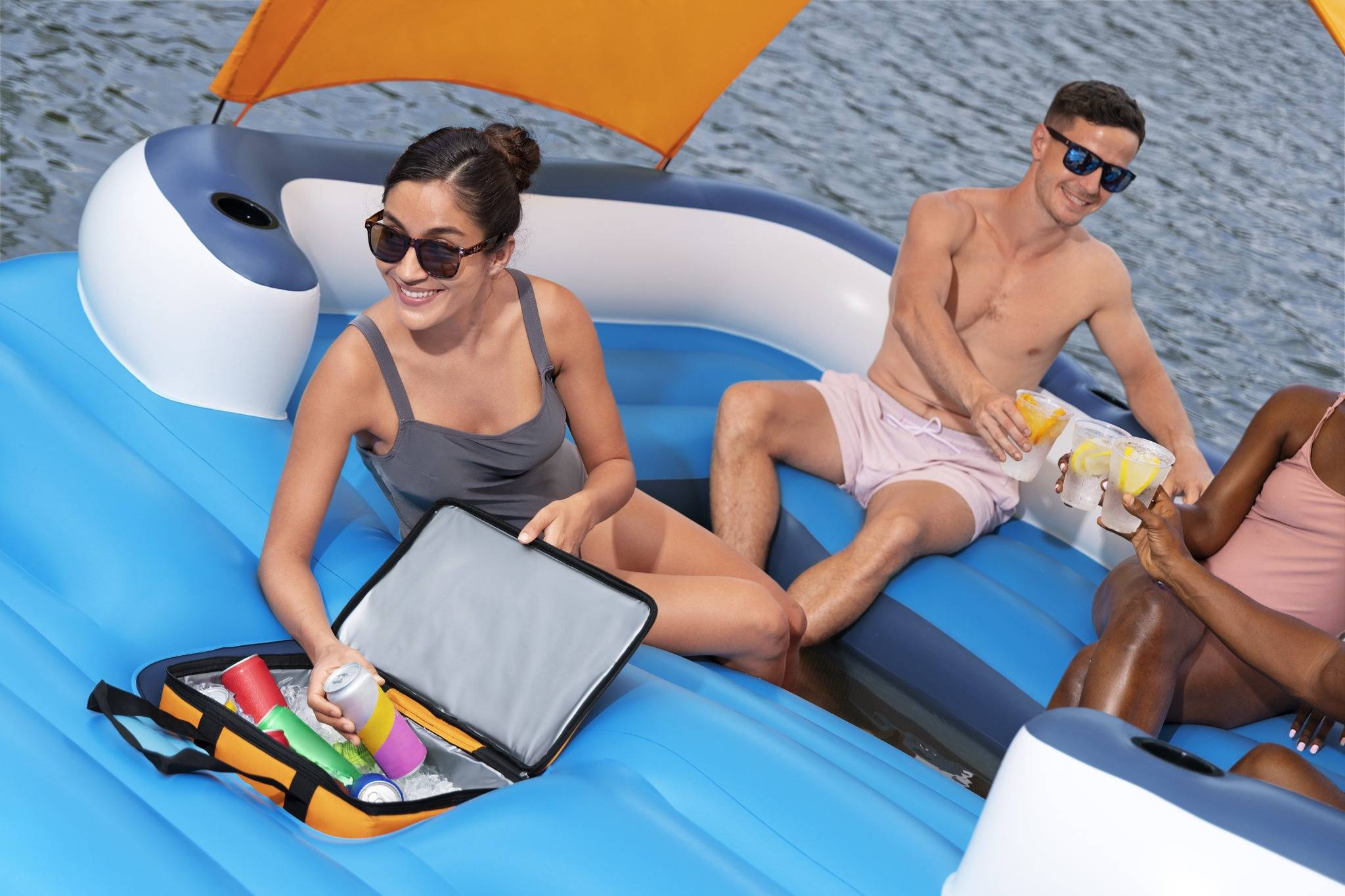 Zwei Personen entspannen sich auf einem aufblasbaren Boot, während sie Getränke halten, neben ihnen steht eine offene Kühlbox mit weiteren Getränken, unter einem sonnigen Himmel.