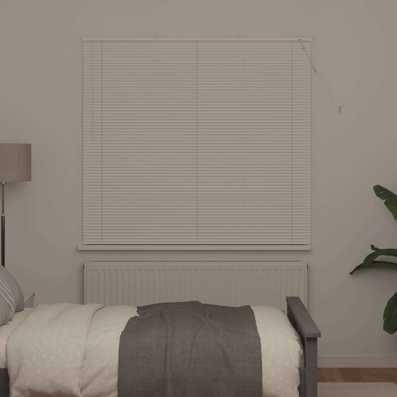 Ein ordentlich gemachtes Bett mit weiß-grauer Bettwäsche steht an einer Wand mit geschlossenen Jalousien. Eine Pflanze ist teilweise auf der rechten Seite sichtbar.