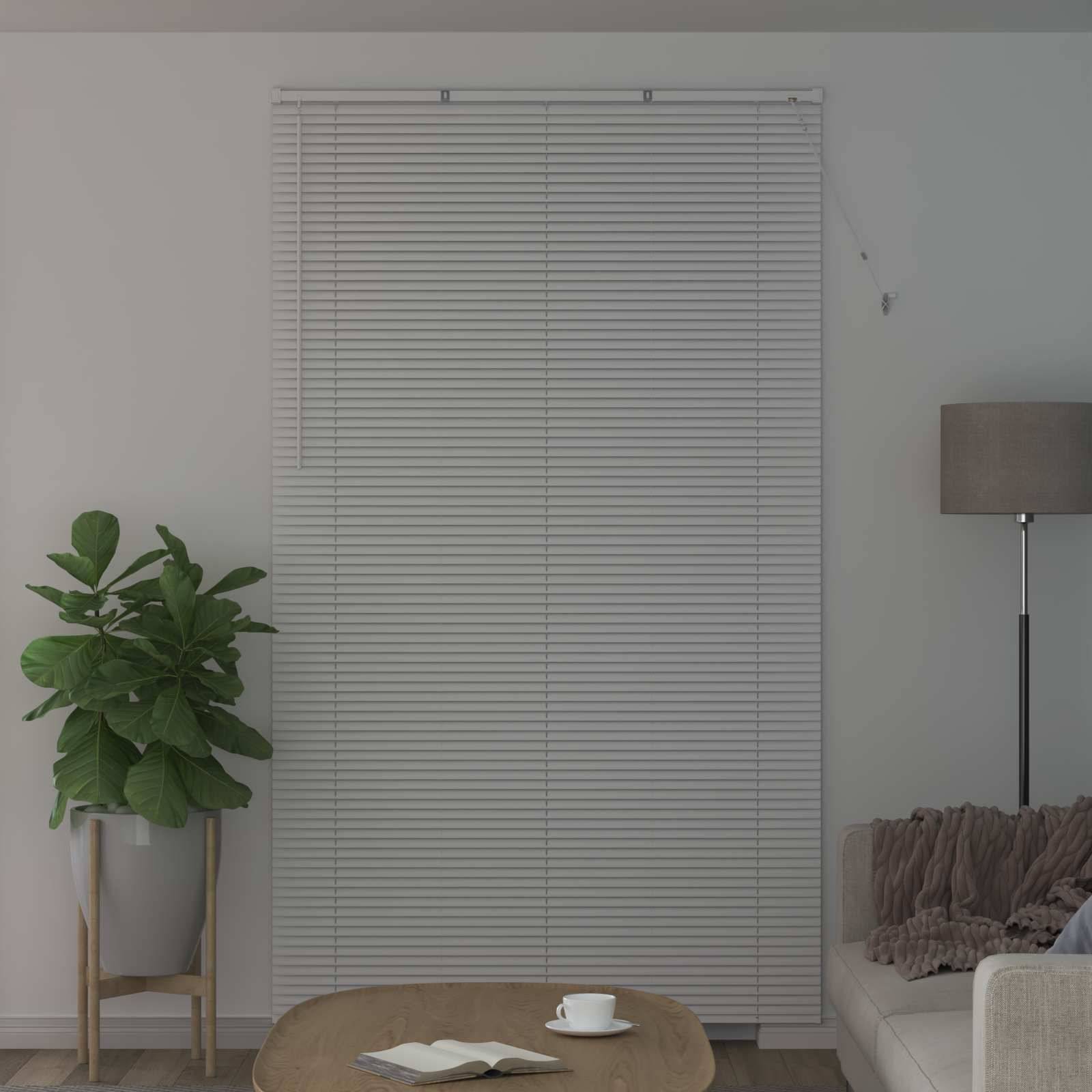 Ein Wohnzimmer verfügt über eine hohe Topfpflanze, geschlossene Jalousien an einem Fenster, eine graue Stehlampe und einen weißen Couchtisch mit einem Buch und einer Tasse.