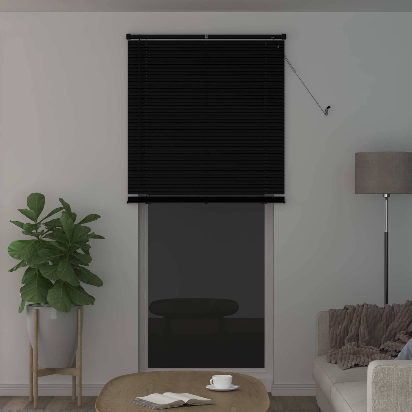 Ein Wohnzimmer mit geschlossenem Fensterrolllo, einer Stehlampe, einer Topfpflanze auf einem Ständer und einem kleinen runden Tisch. Sofa teilweise sichtbar.