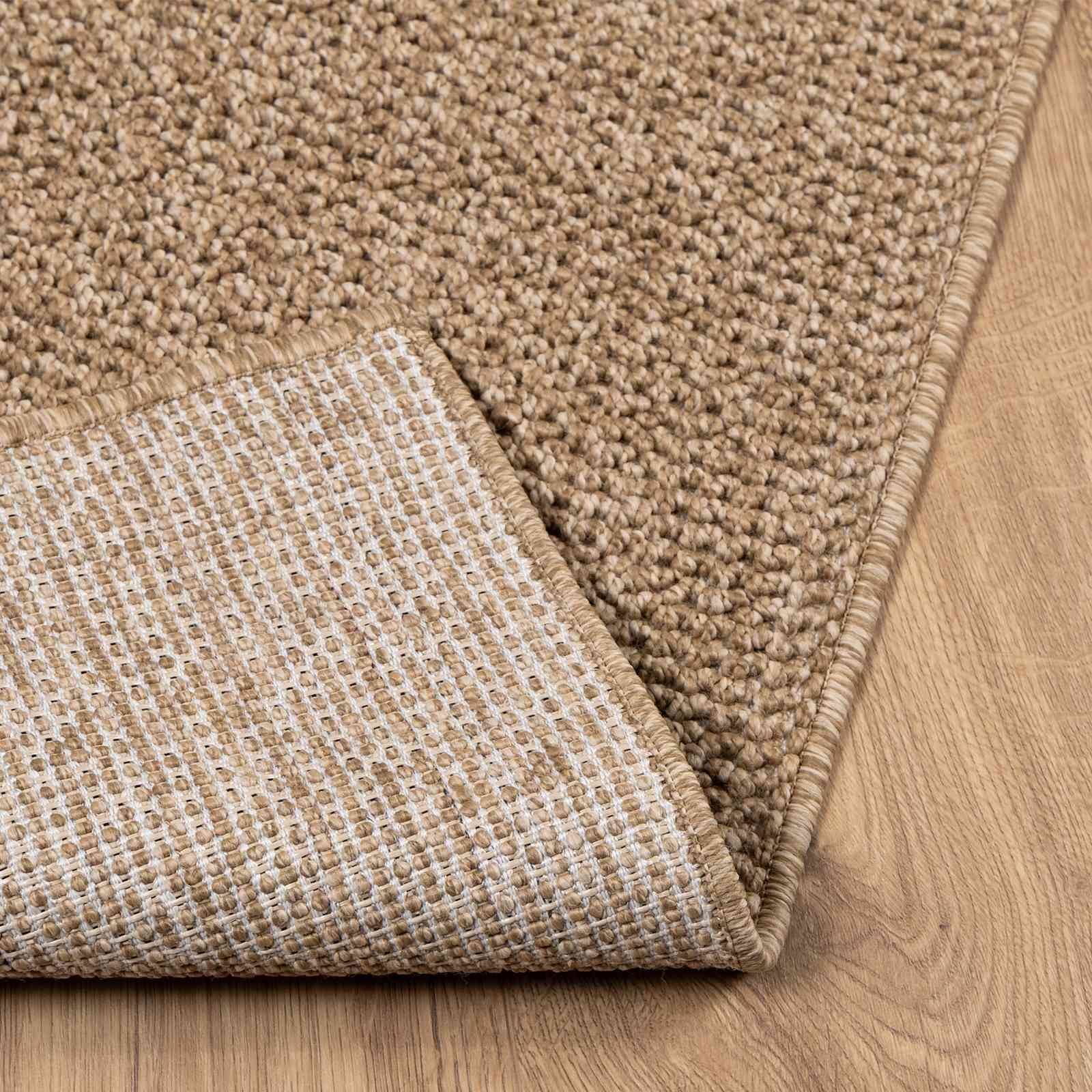 Ein beige gewebter Teppich mit einer sichtbaren Eckenfalte, die eine rutschfeste Rückseite zeigt, auf hellem Holzfußboden gelegt.