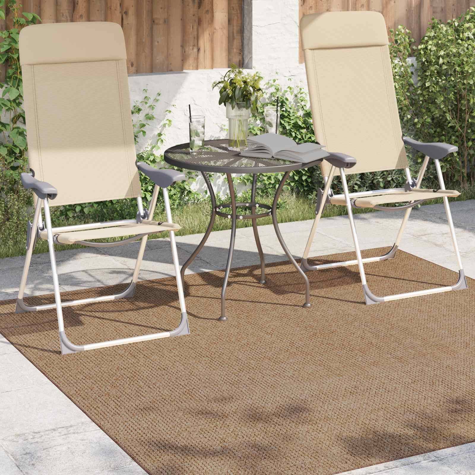 Zwei beige Terrassenstühle neben einem kleinen runden Tisch mit einer Topfpflanze und Büchern, auf einem grauen Teppich angeordnet. Steinboden und Holzwand im Hintergrund.