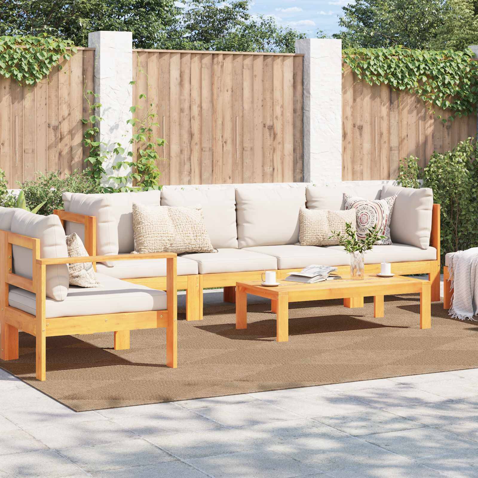 Outdoor-Terrasse mit Holzmöbel-Set, bestehend aus einer Eckgarnitur, einem Tisch und einem Sessel auf einem Teppich. Zaun und Grünflächen im Hintergrund.