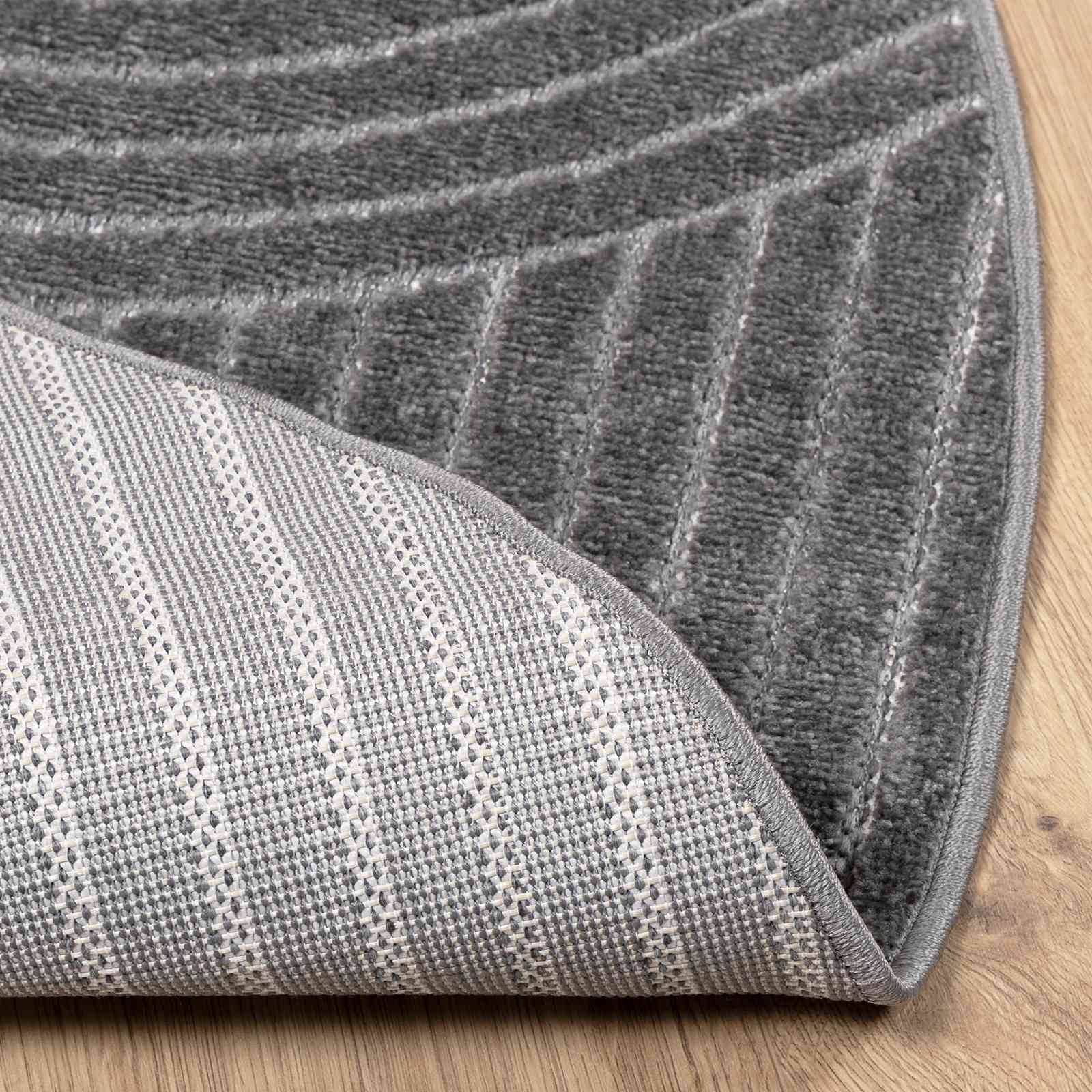 Ein grauer, runder Teppich auf einem Holzboden, der zwei verschiedene Texturen zeigt: ein glattes Streifenmuster oben und eine gitterartige Webstruktur unten.