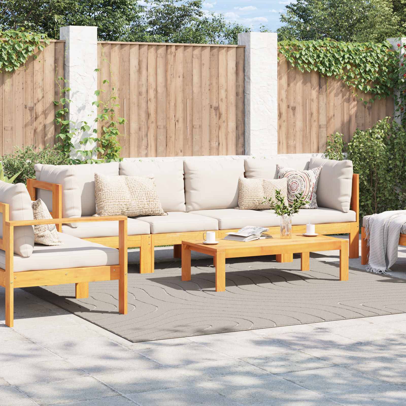 Outdoor-Terrassenmöbel mit hellem Holzrahmen und grauen Polstern, umgeben von einem Holzzaun und Grünflächen. Ein kleiner Couchtisch vervollständigt die Einrichtung.