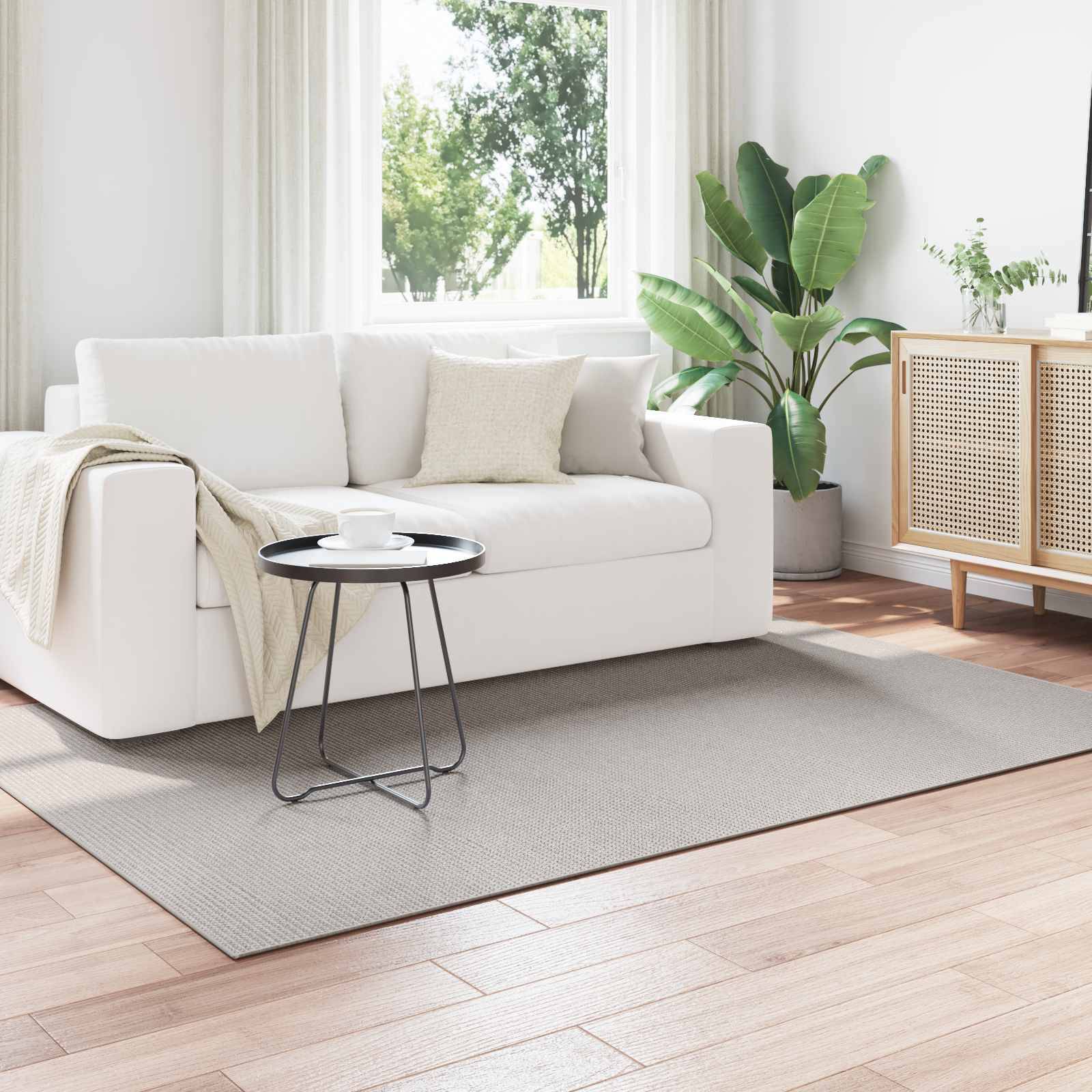 Ein modernes Wohnzimmer mit einem weißen Sofa, einem kleinen schwarzen Beistelltisch, Pflanzen und einem hellholzfarbenen Schrank an einer weißen Wand. Sonnenlicht fällt durch transparente Gardinen.