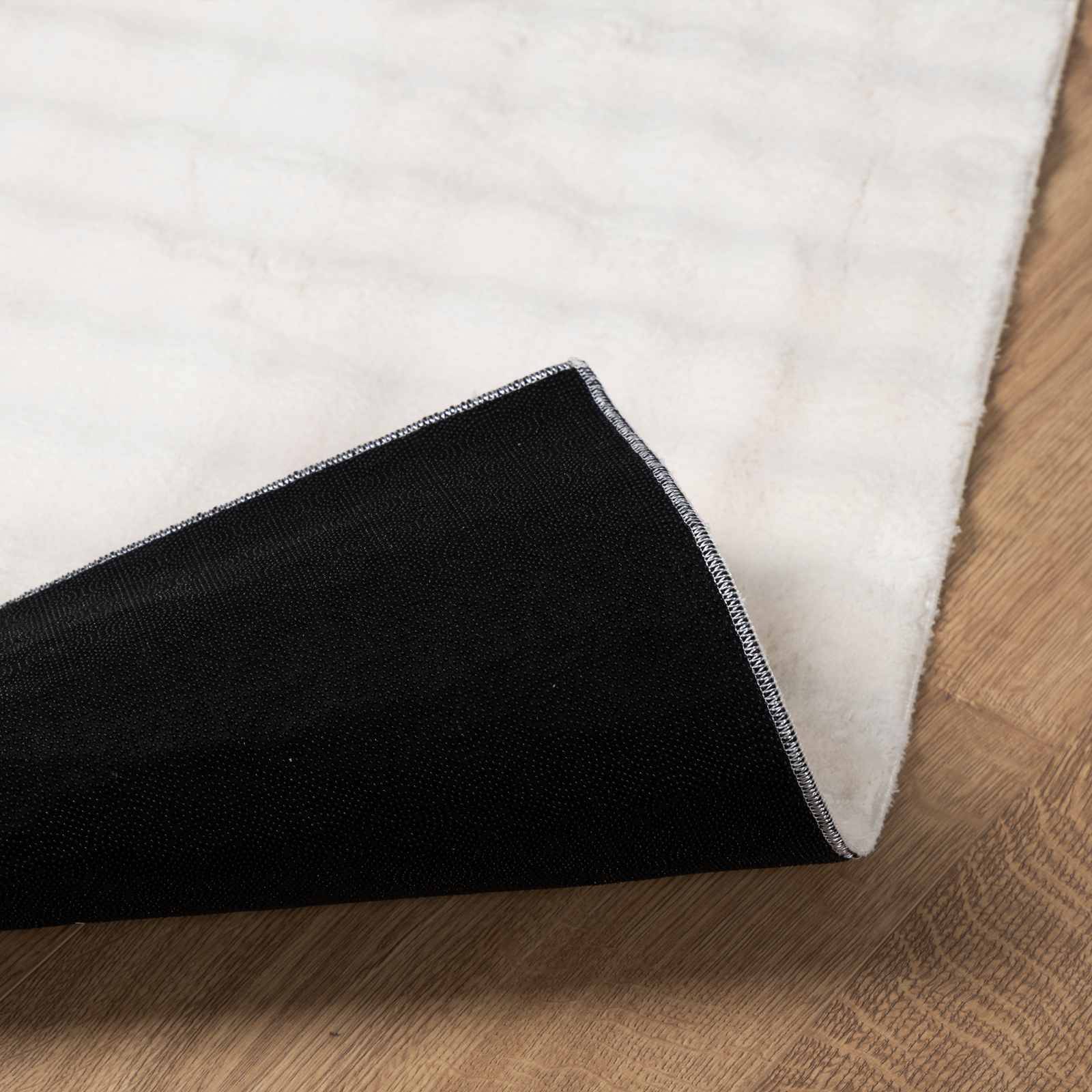 Eine Ecke eines weißen Teppichs ist umgeklappt und zeigt seine schwarze Unterseite, platziert auf einem Holzboden.