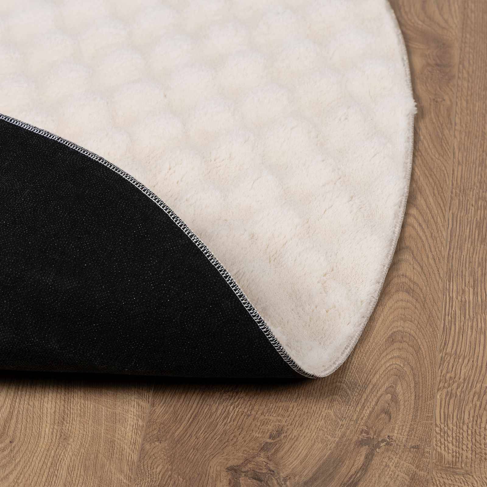 Ein runder, weißer strukturierter Teppich mit einer sichtbar teilweise umgeschlagenen Kante, die eine schwarze rutschfeste Unterseite auf einem Holzboden offenbart.