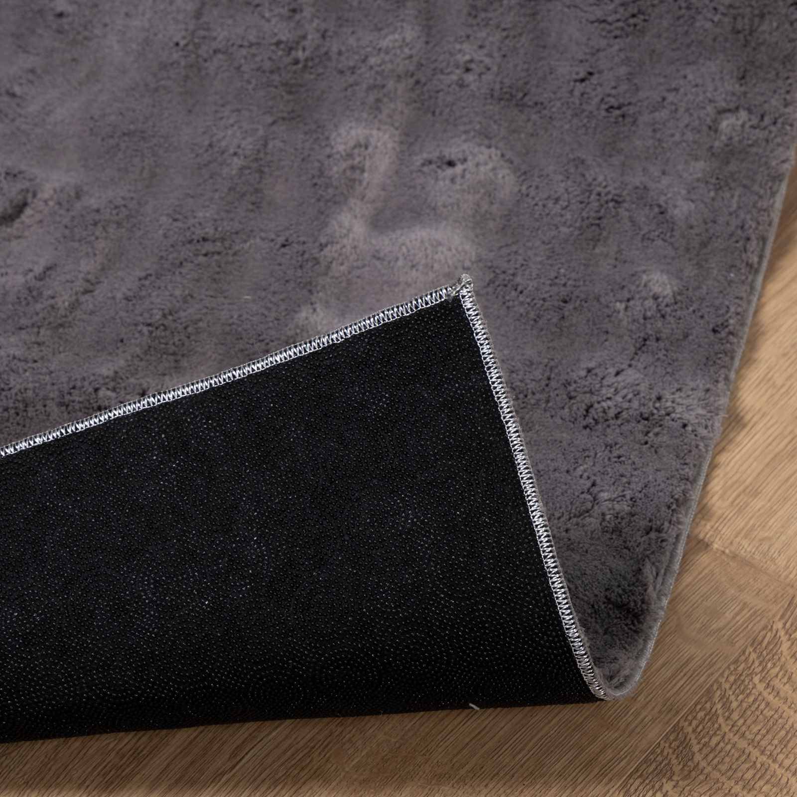 Eine Nahaufnahme eines grauen Teppichs mit schwarzer rutschfester Rückseite, die eine umgeschlagene Ecke zeigt, auf einem Holzboden platziert.