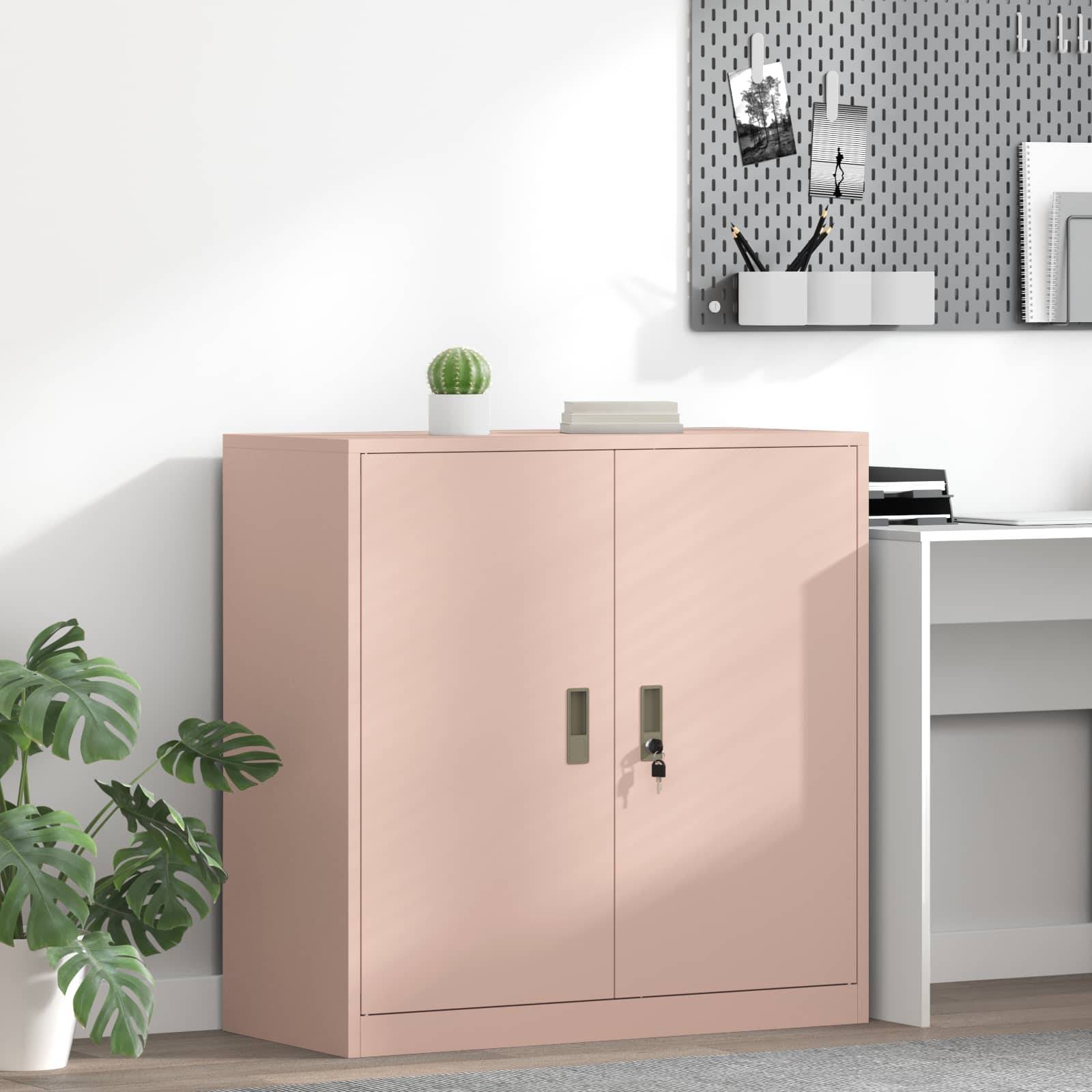 Ein rosa Schrank mit geschlossenen Türen steht neben einem weißen Schreibtisch mit Büromaterial an einer hellen Wand. Ein Pinnwandbrett hängt über dem Schreibtisch.