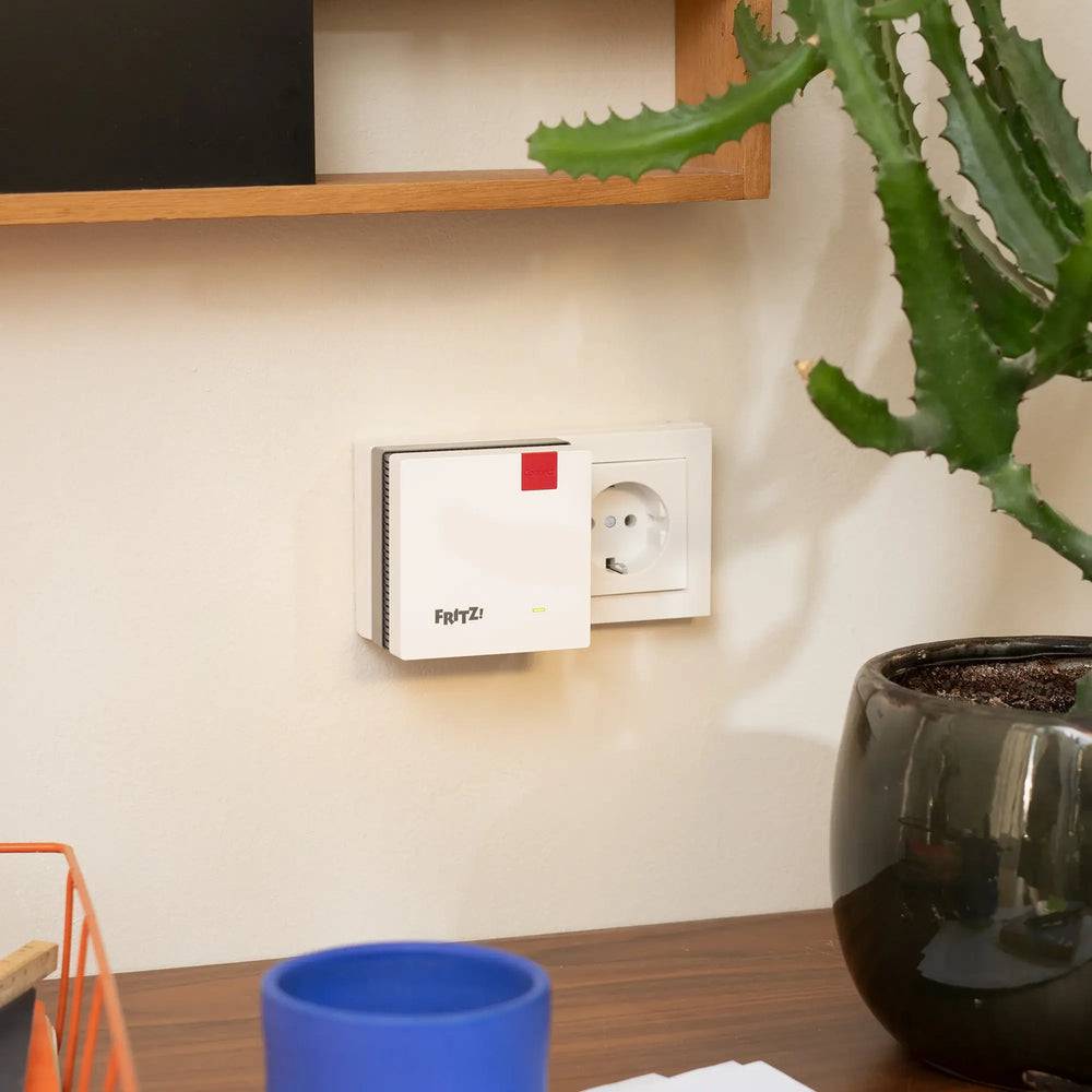 Ein wandmontiertes intelligentes Thermostat neben einer Steckdose in einem Wohnungsinneren, mit einer Kaktuspflanze und Schreibtischgegenständen im Vordergrund.