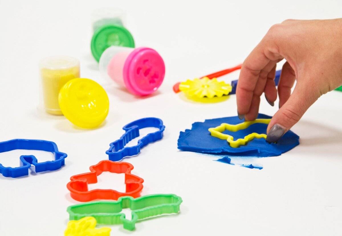 Eine Hand drückt gelbes Knete mit einem sternförmigen Ausstecher. Verschiedene bunte Ausstecher und Knetedosen sind verstreut herum.