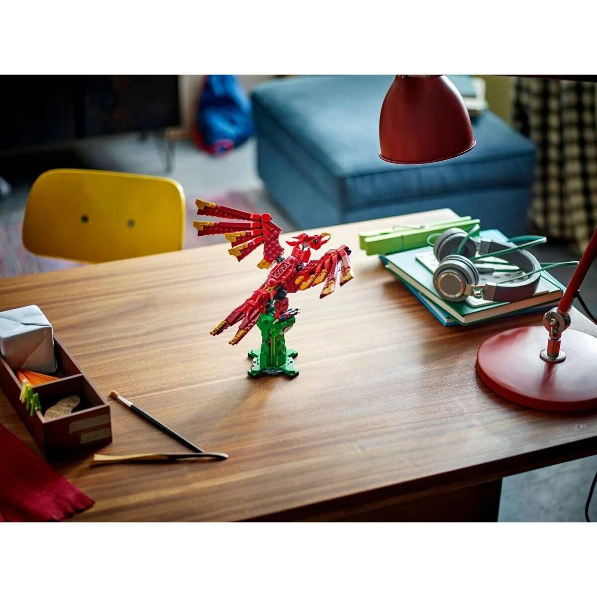 Ein farbenfrohe Vogelmodell aus ineinandergreifenden Bausteinen auf einem Holzschreibtisch, umgeben von einer Lampe, Kopfhörern und Schreibwaren unter warmer Beleuchtung.