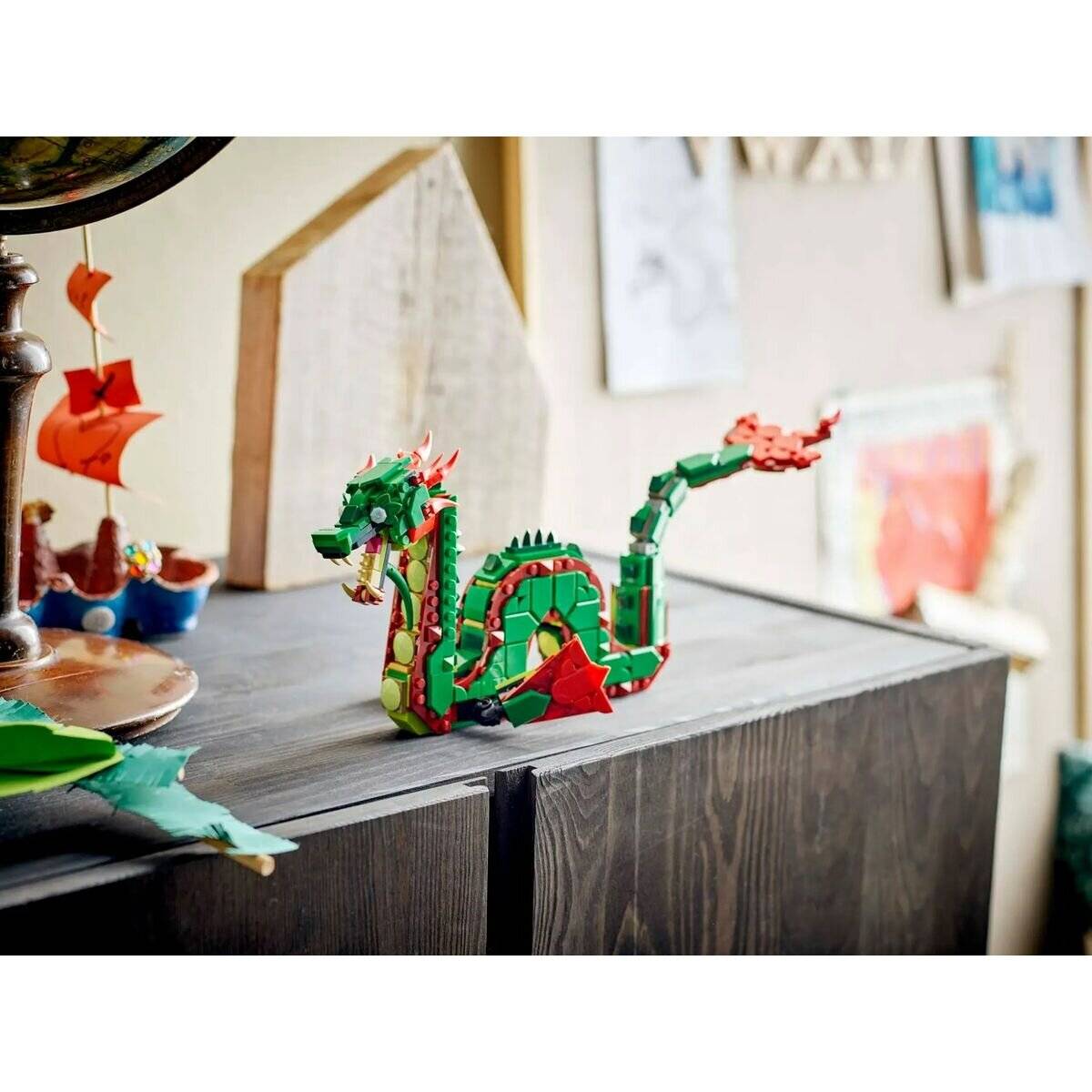 Ein kleines, farbenfroqures Drachenmodell aus Bausteinen sitzt auf einem hölzernen Schrank, umgeben von Kunst- und Bastelmaterialien in einer ungezwungenen Umgebung.