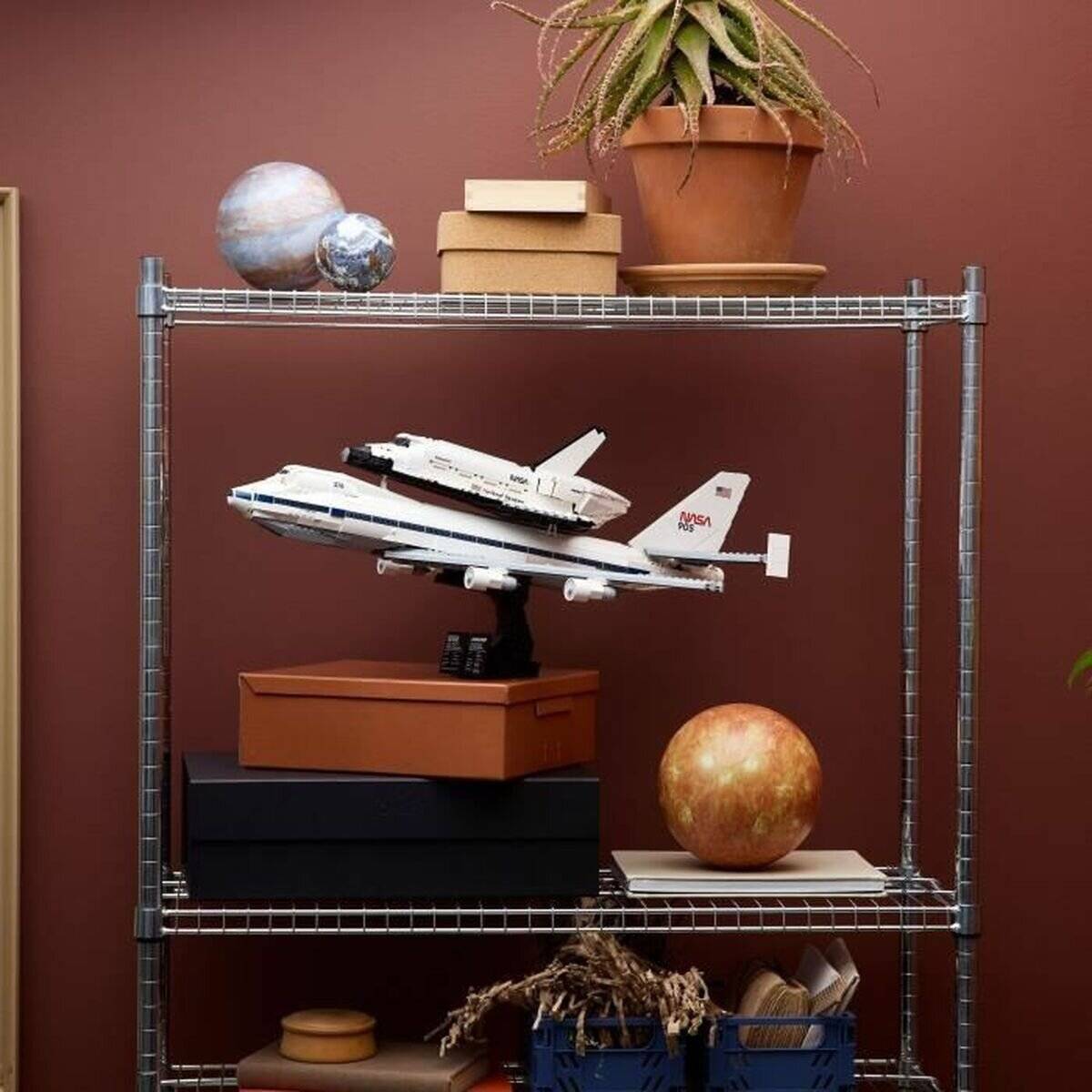 Modellflugzeuge und ein Space Shuttle auf einem Regal ausgestellt, akzentuiert durch Topfpflanzen und Dekorationsgegenstände vor einem braunen Hintergrund.