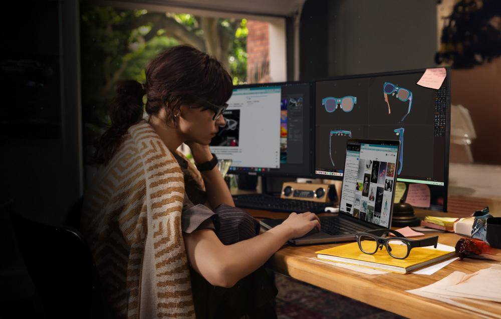 Eine Person, die einen Schal trägt, nutzt eine Computer-Einrichtung mit mehreren Monitoren, die Brillendesigns und Grafikbearbeitungssoftware anzeigen.