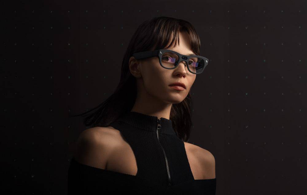 Eine Person, die dunkle, futuristische Brillen und ein schwarzes Reißverschluss-Oberteil vor dunklem Hintergrund trägt und leicht nach oben schaut.