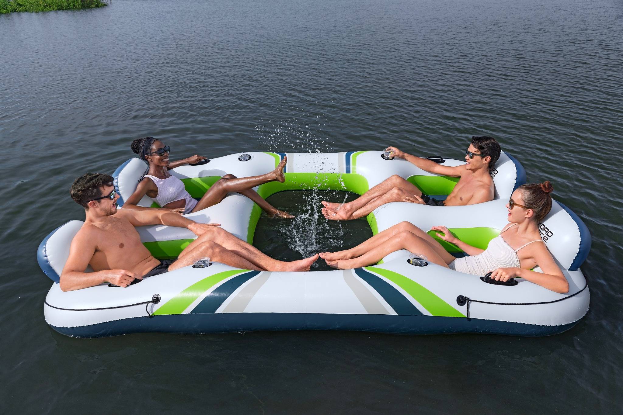 Vier Menschen entspannen sich auf einem aufblasbaren Floß, das auf dem Wasser treibt, lächeln und spritzen sich gegenseitig verspielt in der Sonne.