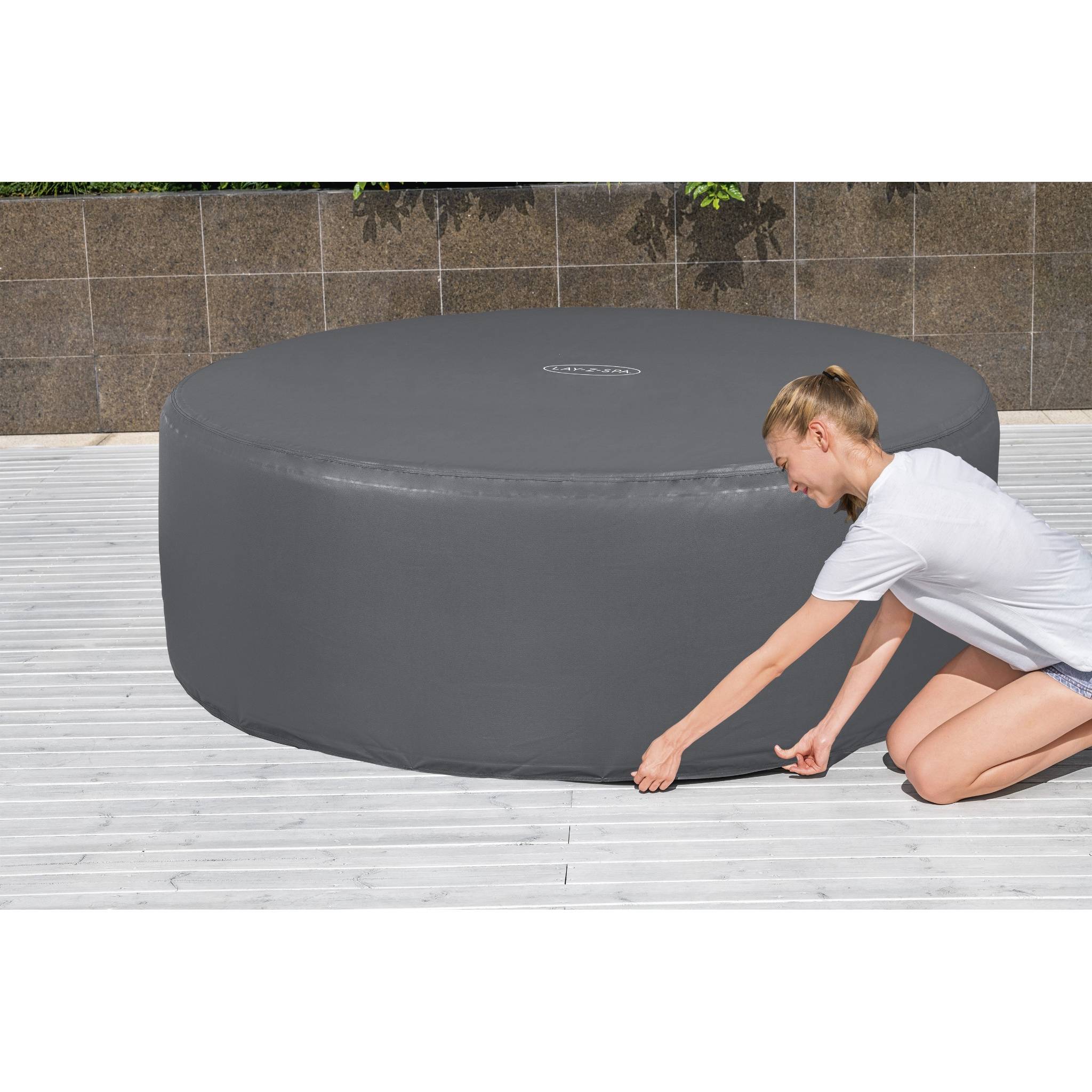 Eine Person kniet auf einem Holzdeck und befestigt eine runde Schutzabdeckung über einem Außen-Whirlpool. Im Hintergrund eine braun geflieste Wand.