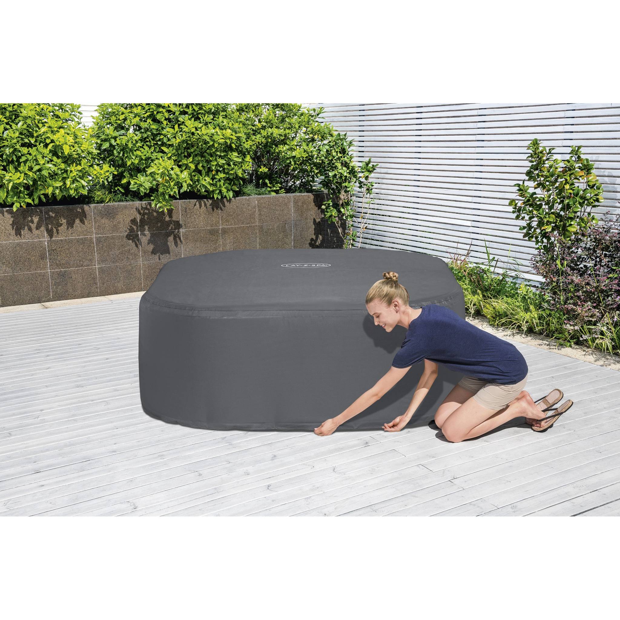 Eine Person befestigt eine dunkle Abdeckung über einem großen, freistehenden Whirlpool auf einer Holzterrasse mit grünen Pflanzen und einem Zaun im Hintergrund.