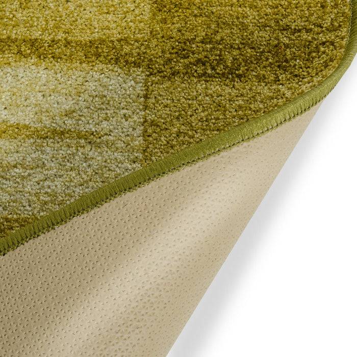 Ein strukturierter, grüntöniger Teppich mit rutschfester Unterseite, der ein Farbverlaufsmuster aus Quadraten zeigt. Die Ecke ist leicht angehoben, um die Unterseite freizulegen.