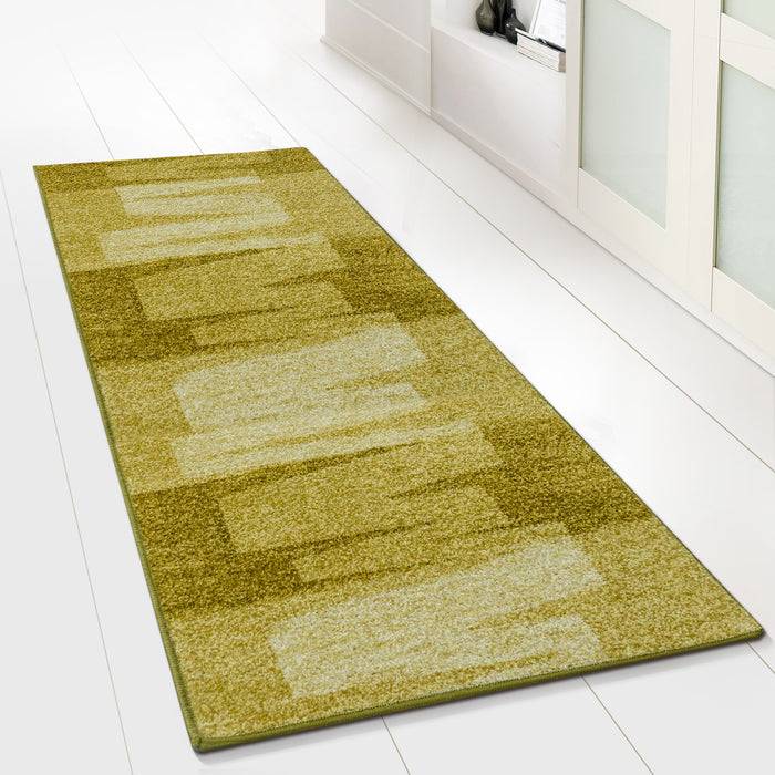 Ein langer, rechteckiger Teppich mit grünem und beigem Muster liegt auf einem weißen Boden neben einem weißen Bücherregal, das mit verschiedenen Gegenständen gefüllt ist.