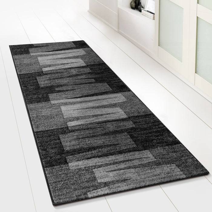 Ein grauer rechteckiger Teppich mit geometrischem Muster liegt auf einem weißen Fliesenboden neben einem weißen Schrank.