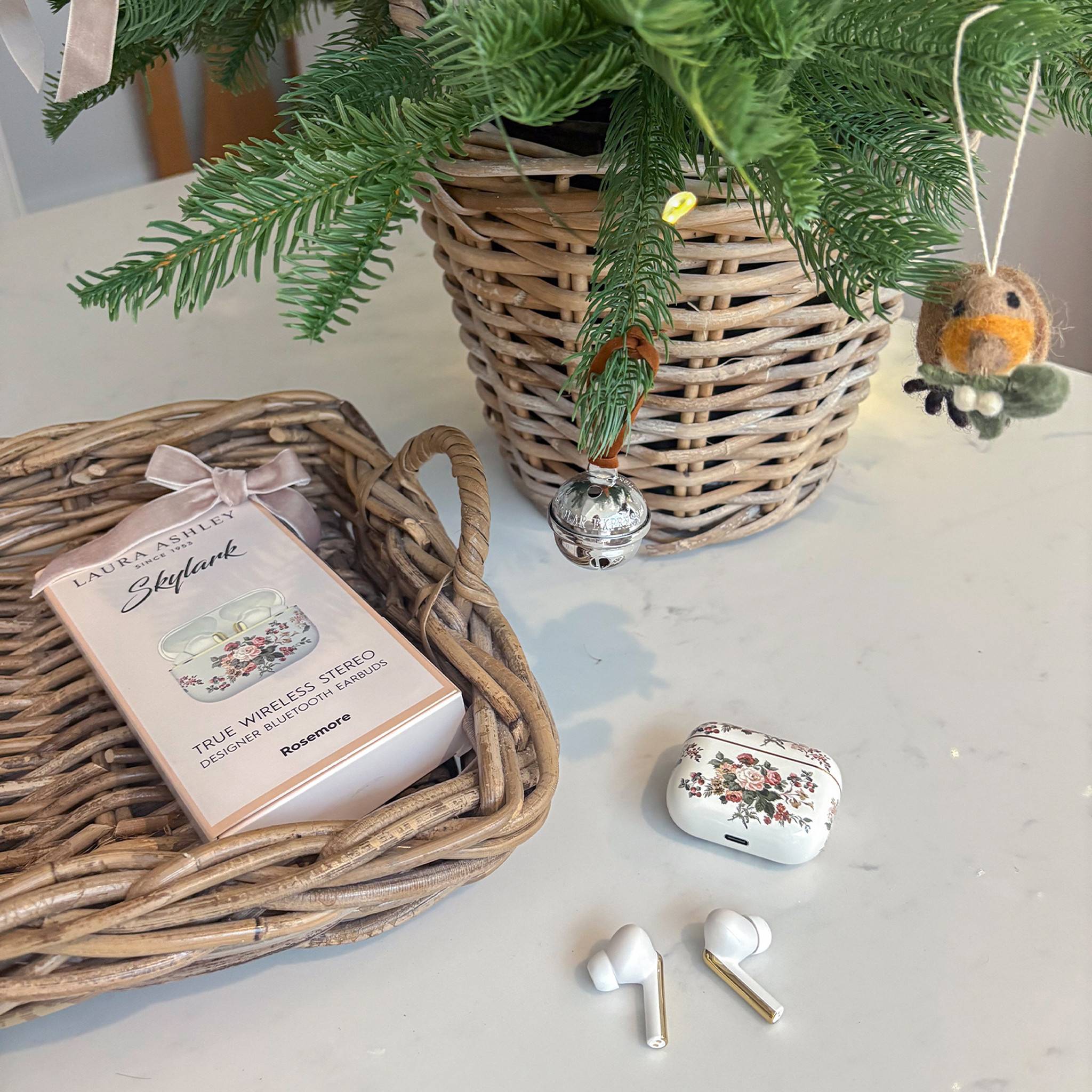 Ein Korb mit einer Topfpflanze und festlichen Dekorationen steht auf einem Tisch. In der Nähe befinden sich kabellose Ohrhörer, ihr Etui mit floralem Design und ein Tablett mit einer Box mit der Aufschrift „True Wireless Stereo