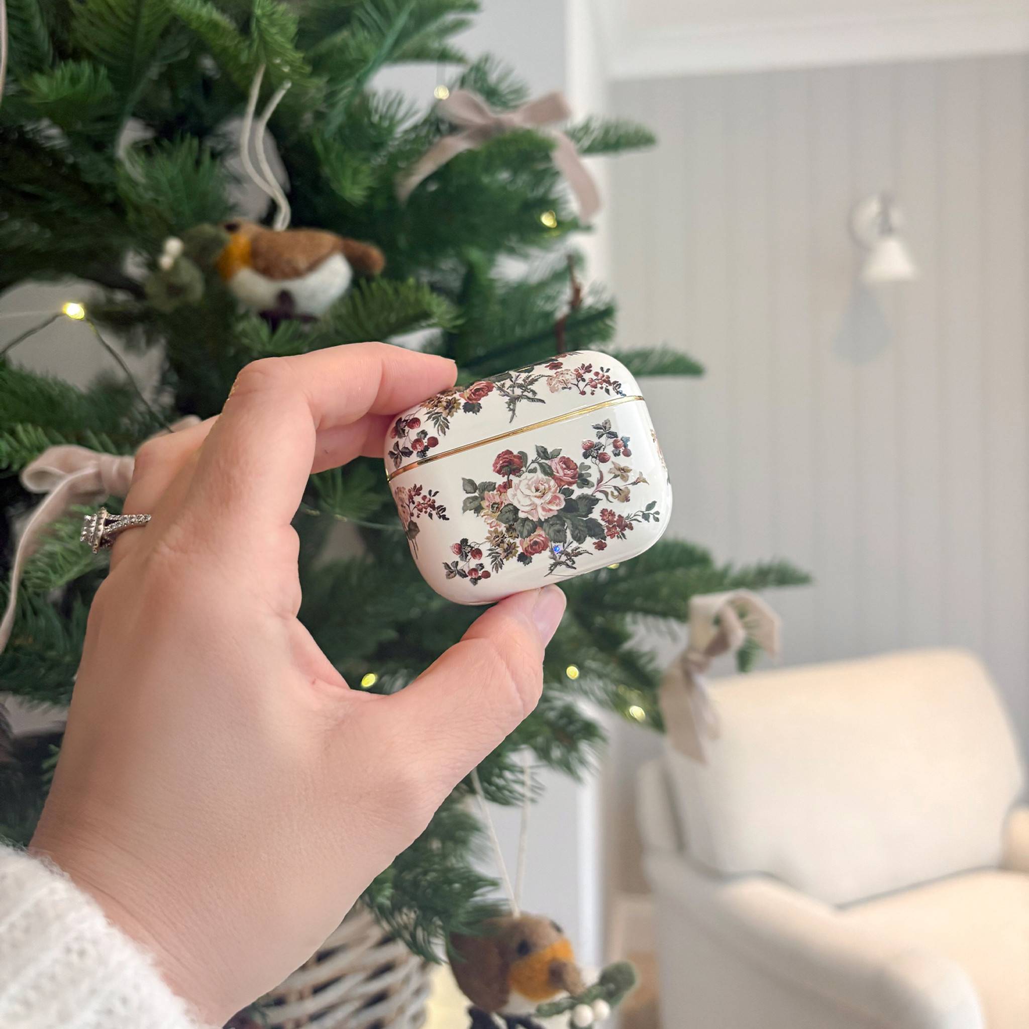 Eine Hand hält ein Etui für kabellose Ohrhörer mit Blumenmuster vor einem geschmückten Weihnachtsbaum, mit einem weißen Stuhl im Hintergrund.