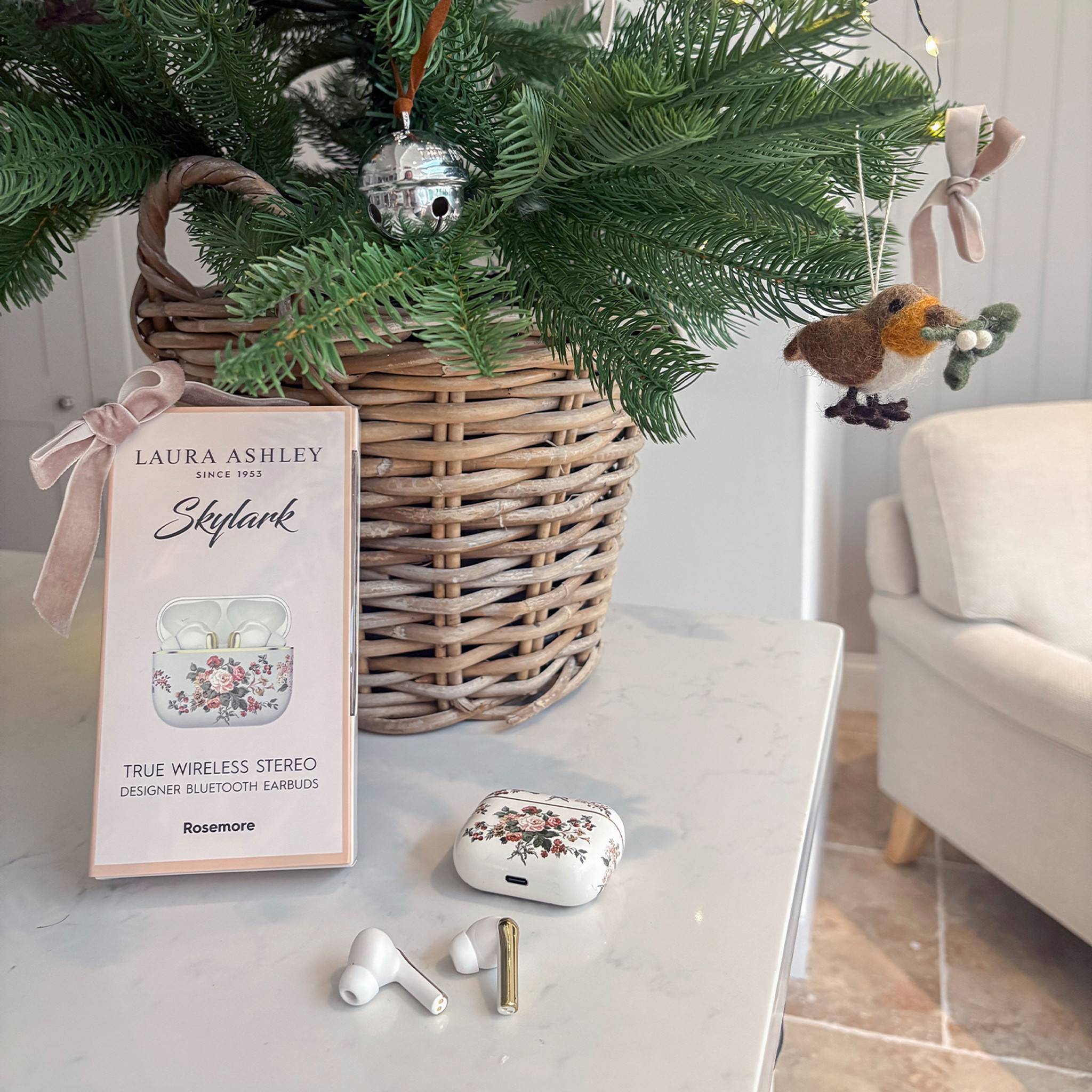 Ein Weidenkorb mit einem dekorierten Tannenzweig steht auf einem Tisch neben einer Schachtel und einem Paar kabellose Ohrhörer mit Blumenmuster, die mit 