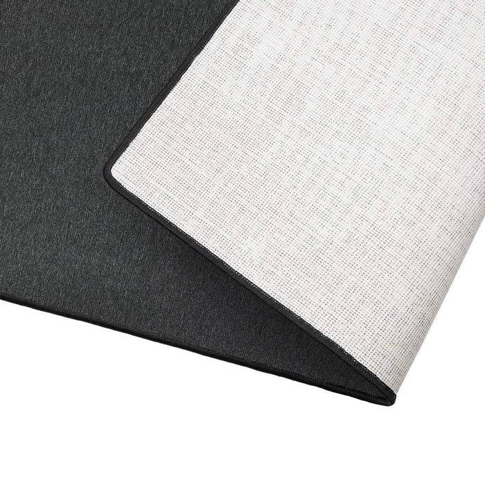 Eine Nahaufnahme einer hellgrauen Teppichkante, nach oben umgeklappt, um die rutschfeste Rückseite zu zeigen, mit sichtbarer Textur und Polsterstärke.