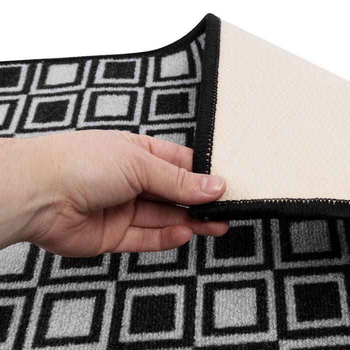 Eine Hand hebt die Ecke eines grau-schwarzen geometrisch gemusterten Teppichs an und zeigt seine rutschfeste Rückseite.