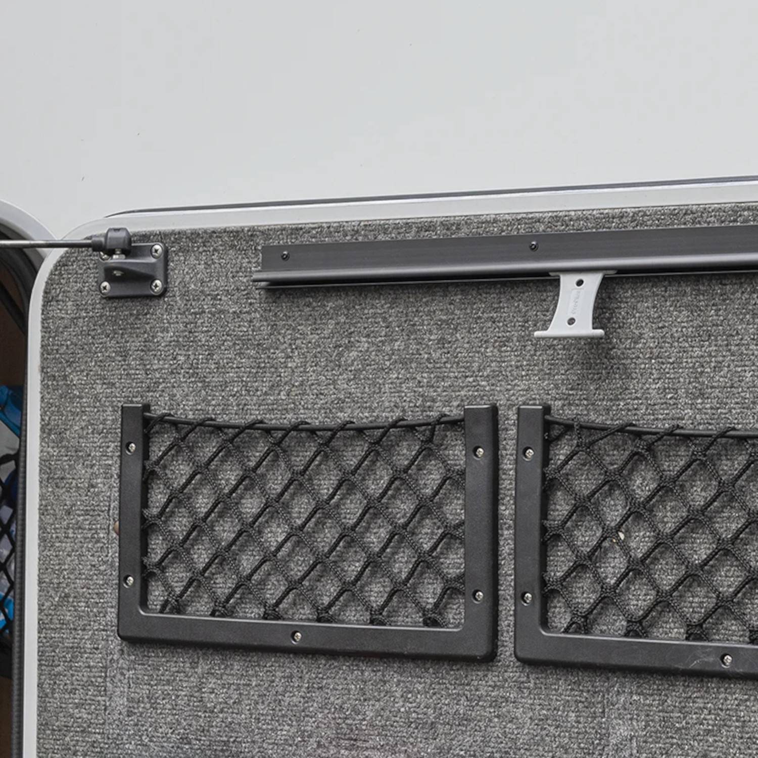 Eine graue, teppichbelegte Wand mit zwei schwarzen Netzfächern, die daran montiert sind, wahrscheinlich zur Aufbewahrung. Ein silberner Haken befindet sich über den Fächern.