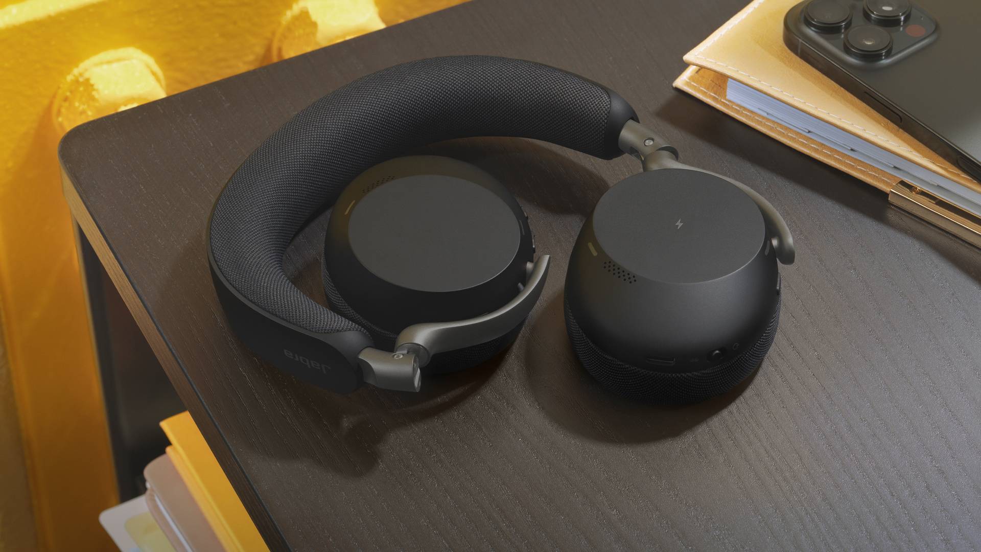 Schwarze Over-Ear-Kopfhörer ruhen auf einem dunklen Holzschreibtisch neben einem Notizbuch und einem Telefon.