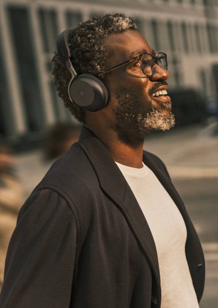 Ein lächelnder Mann mit Brille und Over-Ear-Kopfhörern schaut nach oben im Freien vor einem Gebäude.