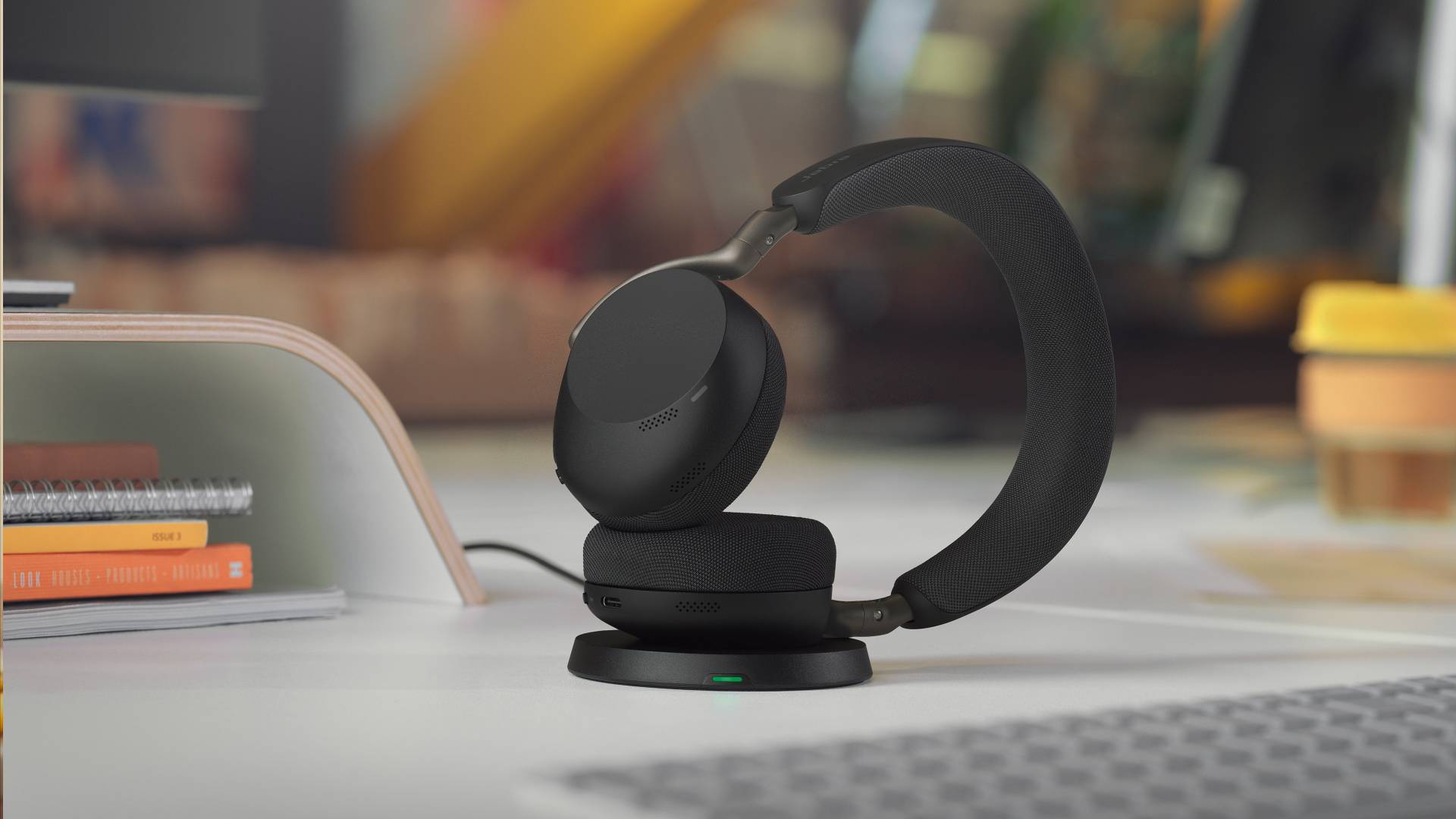 Schwarze Over-Ear-Kopfhörer ruhen auf einem Ladegerät auf einem Schreibtisch, mit einem Laptop im Vordergrund und Büchern daneben.
