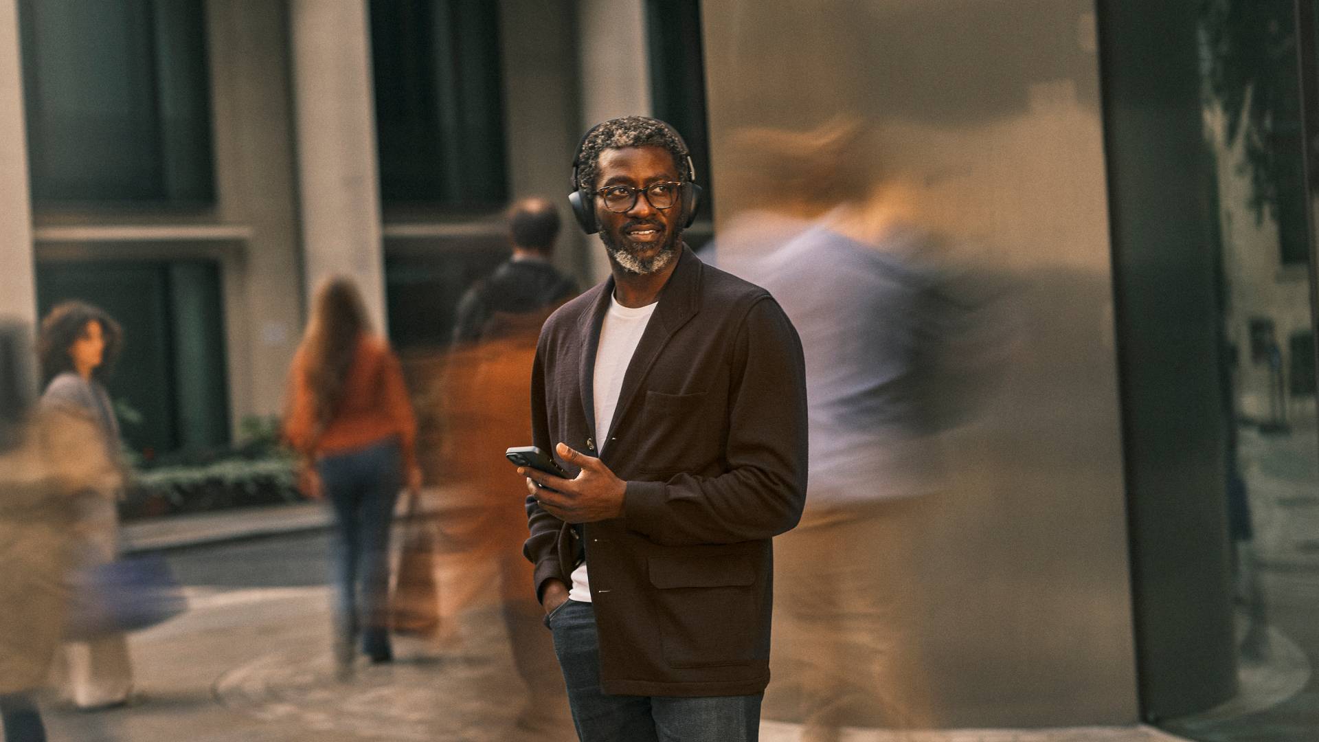 Lächelnder Mann mit einem Telefon in der Hand steht auf einem Gehweg in der Stadt, mit unscharfen Fußgängern und Gebäudespiegelungen hinter ihm.