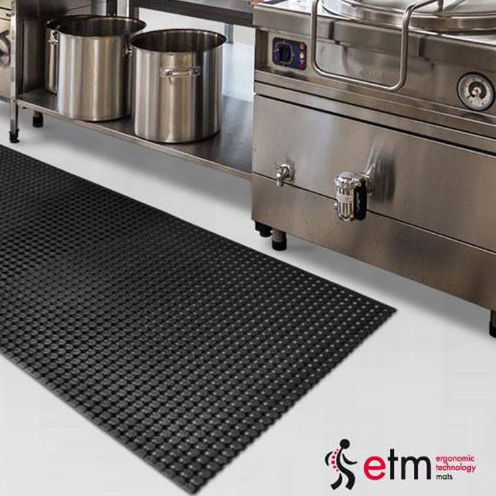 Gewerbliche Küche mit großen Töpfen auf einer Edelstahlarbeitsplatte, neben industriellen Kochgeräten. Eine strukturierte schwarze Matte liegt auf dem Boden davor.