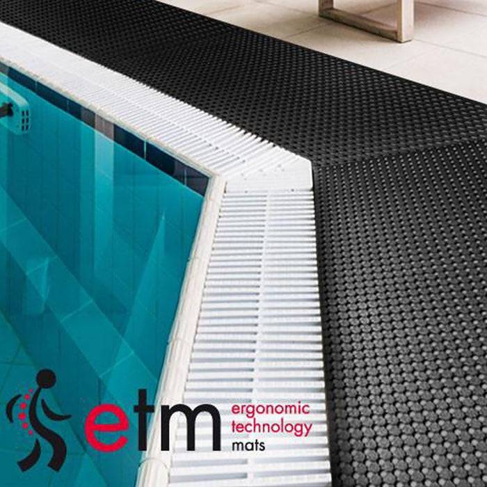 Eine schwarze ergonomische Matte mit strukturierter Oberfläche liegt neben einem Hallenbad und zeigt das Logo mit dem Text: ergonomic technology mats.